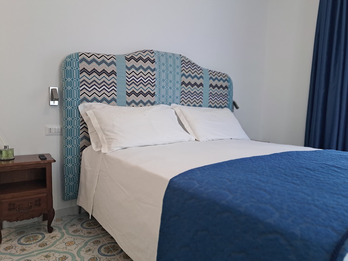 A spacious bed is highlighted by a patterned headboard in shades of blue and white. White linens are paired with a deep blue bedspread. A wooden nightstand is positioned beside the bed, featuring a small decorative item. A dark curtain covers a window, allowing for privacy.