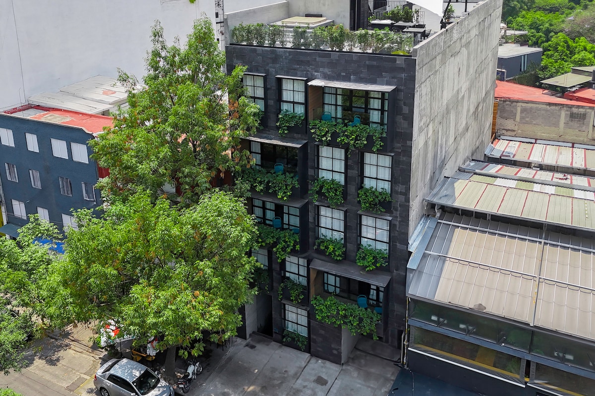 A modern building is showcased, featuring a façade adorned with greenery and large windows. A sprawling tree provides shade at the entrance, enhancing the lively exterior. The surrounding area includes additional structures with varied architecture, contributing to the neighborhood's urban appeal.
