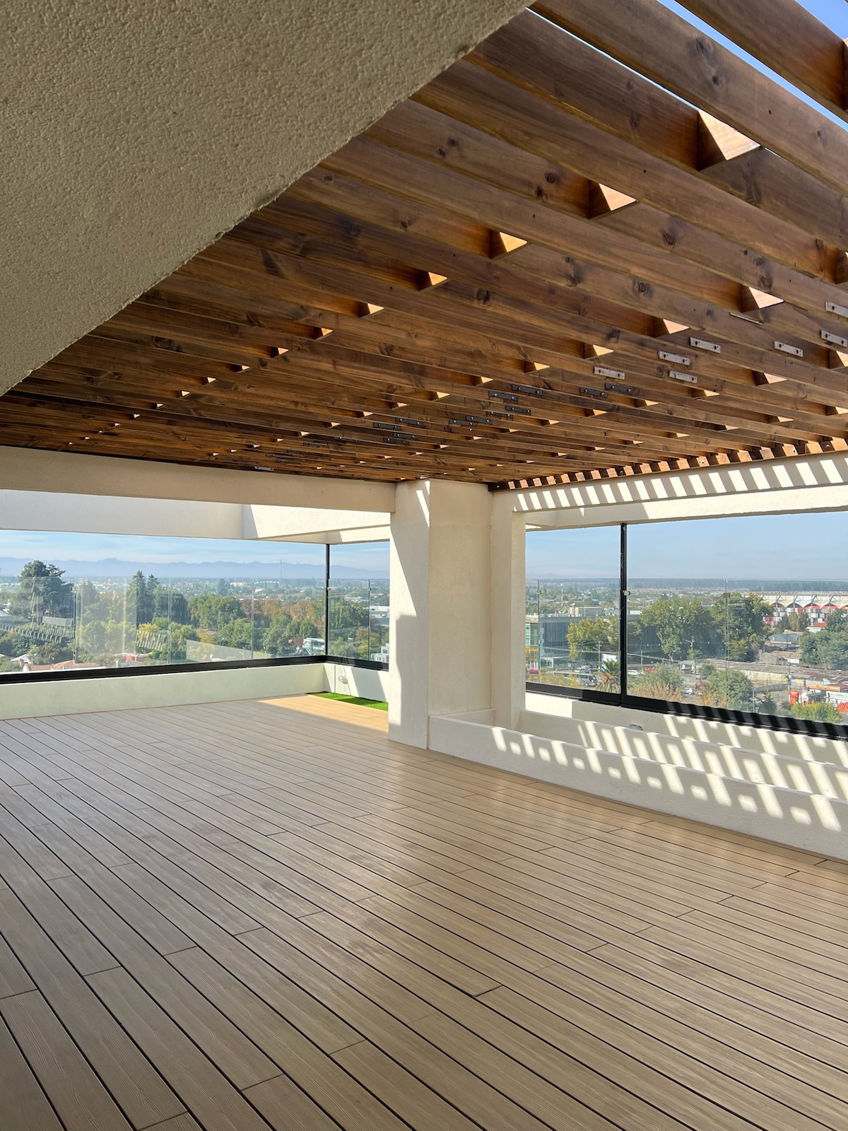 An expansive outdoor area is displayed with wooden slats overhead, allowing natural light to filter through. Large glass windows provide panoramic views of the surrounding landscape, while the wooden flooring adds warmth to the space. The area invites relaxation and offers a serene atmosphere.