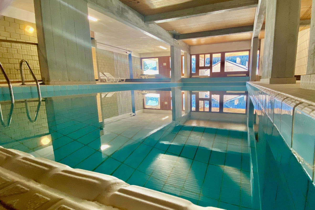 An indoor heated pool exhibits calm blue water, reflecting the surrounding light and architecture. Large windows provide views of the outdoors, while a few lounge chairs are arranged nearby for relaxation.