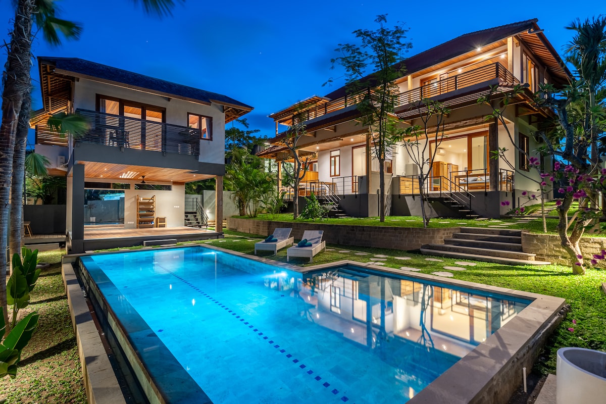 An inviting view of a well-lit pool area is framed by two spacious buildings, surrounded by lush greenery. The pool is illuminated, reflecting the evening sky, with two daybeds positioned nearby for relaxation. Pathways lead through landscaped gardens, creating a serene outdoor atmosphere.