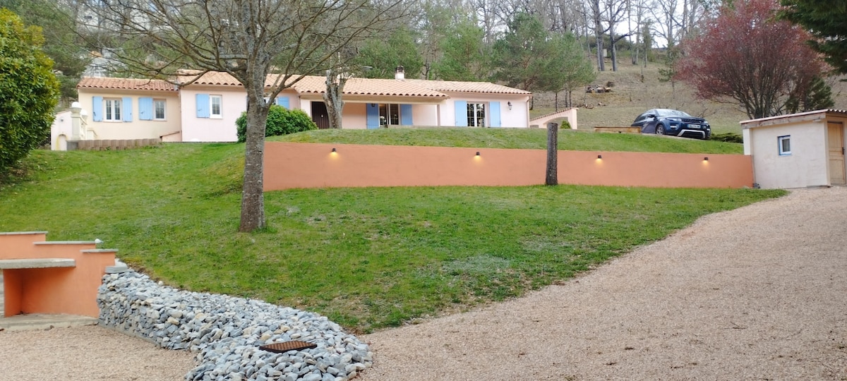 The exterior of a spacious house is showcased, featuring a gently sloping lawn with trees and shrubs. Soft lighting along the wall highlights the structure, while a gravel path leads to the entrance. A vehicle is parked nearby, adding to the property’s accessibility.