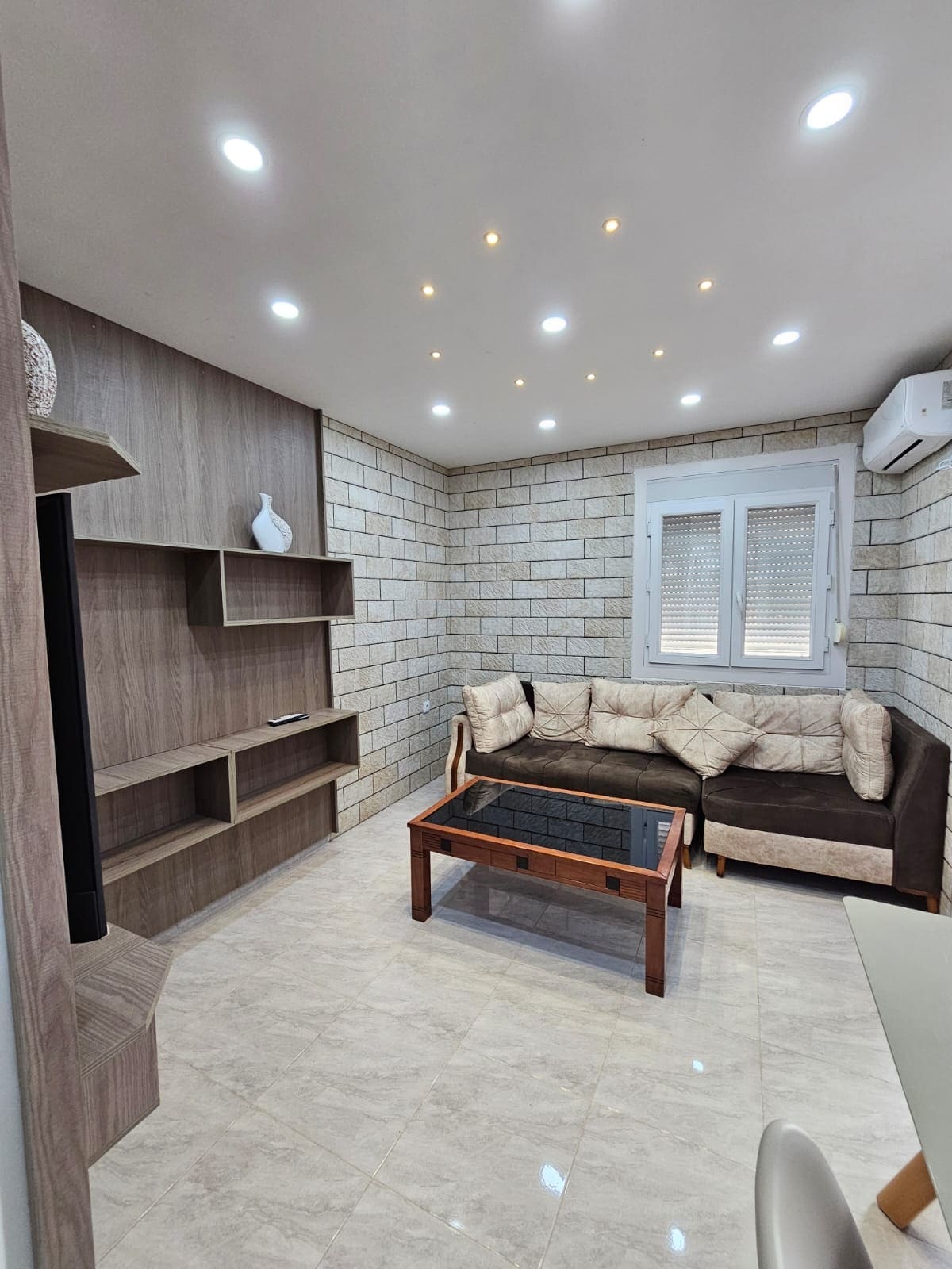 A modern living space is shown, featuring a dark sofa with light-colored cushions and a wooden coffee table. The walls are adorned with light-colored bricks, and built-in shelving is visible on one side. Soft lighting is provided by recessed ceiling lights.