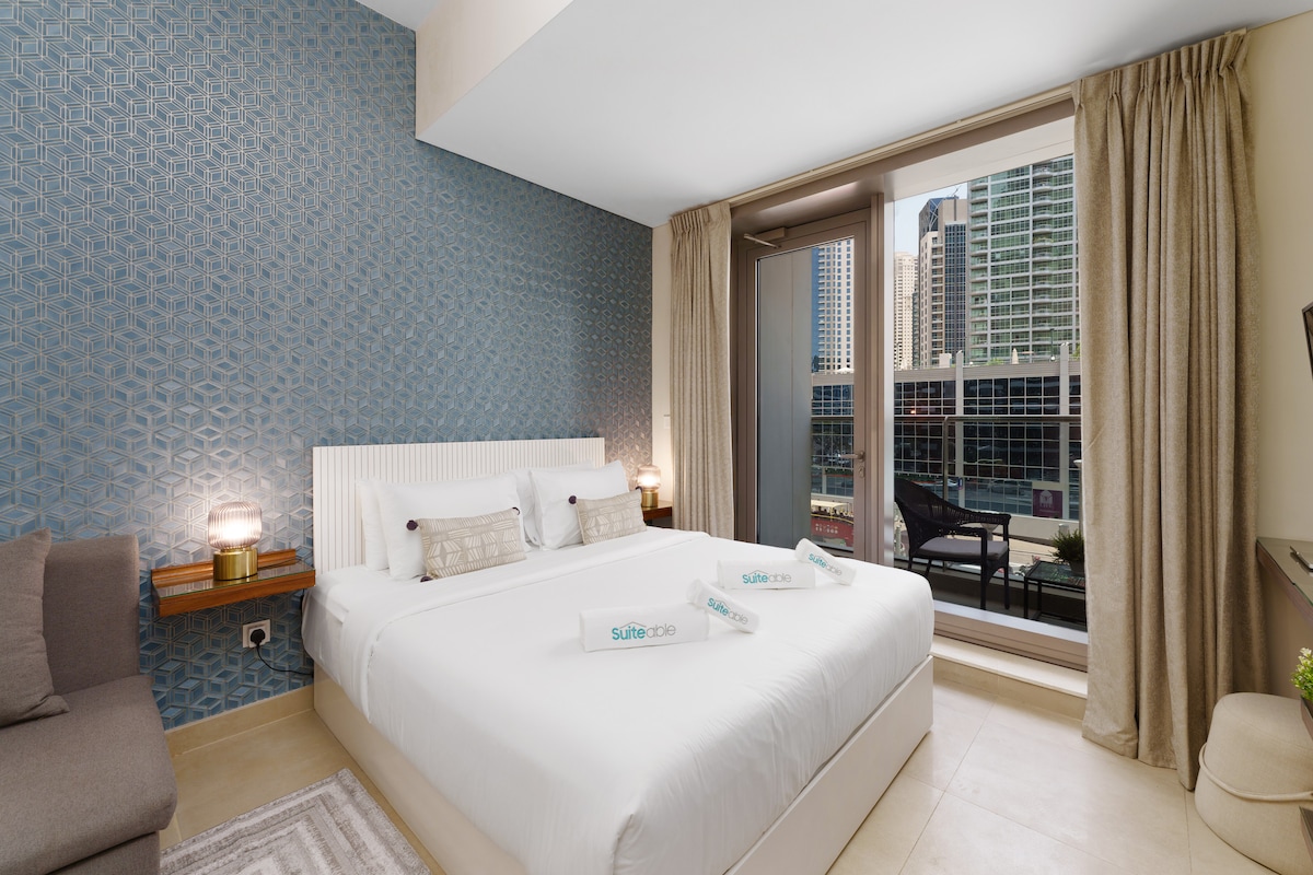 A spacious sleeping area features a large bed with soft linens and two decorative pillows. Natural light enters through a large window framed by light curtains, offering a view of the city skyline. A cozy seating area with a sofa complements the modern interior.