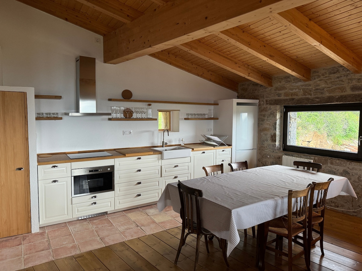 A spacious kitchen features wooden ceilings and stone walls, with a long dining table surrounded by several chairs. Modern appliances, including an oven and refrigerator, are integrated into the cabinetry, while a window allows natural light to brighten the area.