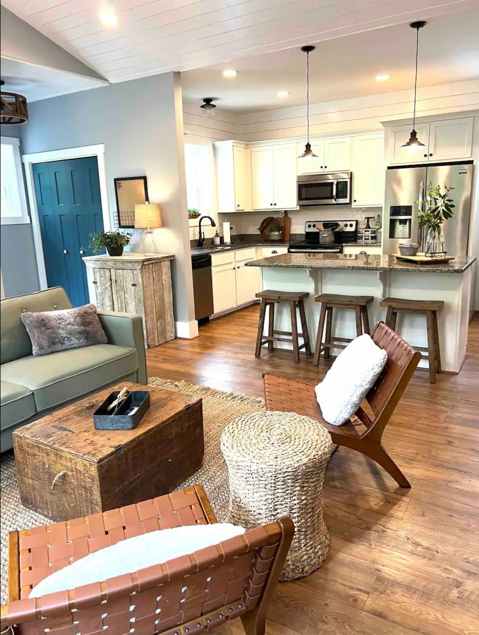 An open living area features a spacious couch with a textured pillow and a wooden coffee table. Two woven chairs flank the coffee table, with the kitchen visible in the background. Modern appliances and light cabinetry are present, illuminated by pendant lighting.