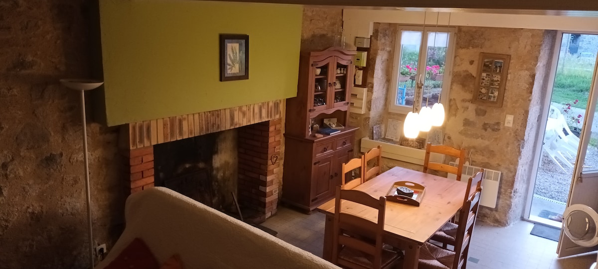 A rustic dining area features a wooden table surrounded by simple chairs, with a large stone fireplace providing a cozy focal point. A bright green accent wall contrasts with the natural stone walls. Soft lighting is emitted from a cluster of hanging fixtures above the table.