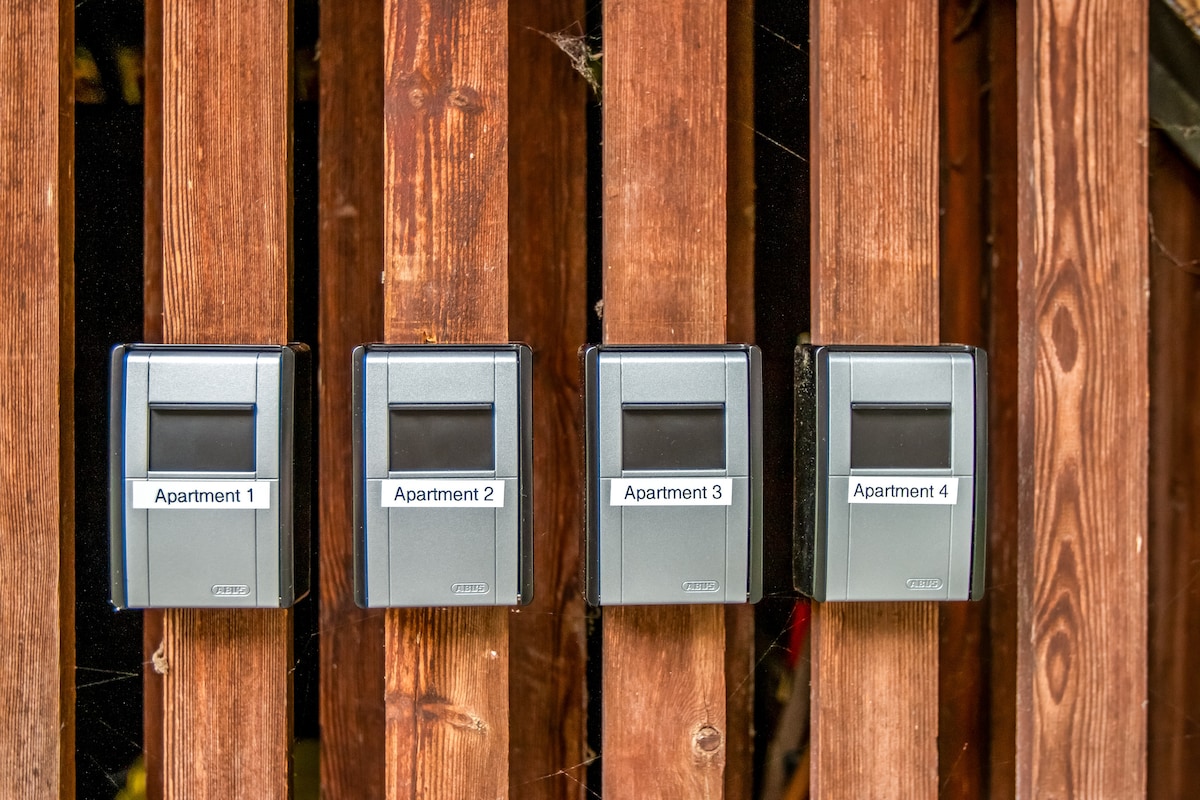 Four mailboxes are mounted on a wooden fence, each labeled with the corresponding apartment number. The boxes feature a sleek silver design, contrasting with the rich brown of the surrounding wood.