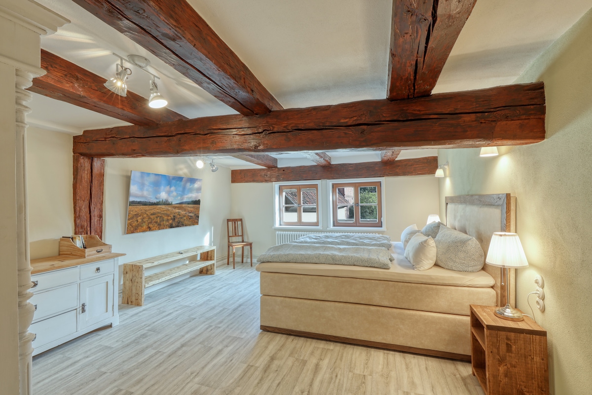A spacious room is featured, highlighting wooden beams across the ceiling and a large window providing natural light. A comfortable bed with soft bedding is positioned against the wall, complemented by bedside tables with lamps. A simple white dresser and a seating area are also visible.