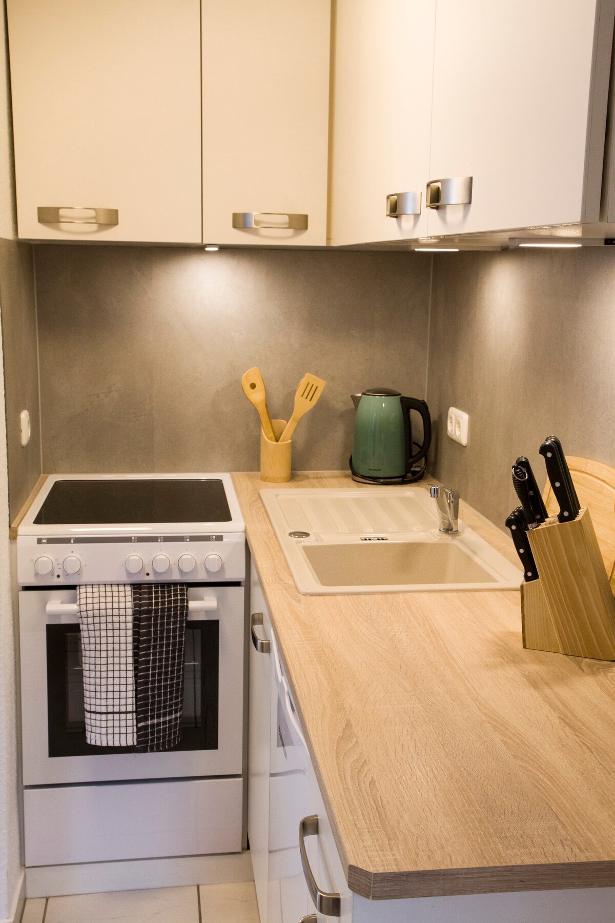 The kitchen features modern cabinetry and sleek surfaces. Essential appliances include a stove, kettle, and a sink. Cooking utensils are organized neatly, and a knife block is prominently displayed on the countertop. Soft lighting enhances the functional and welcoming atmosphere.