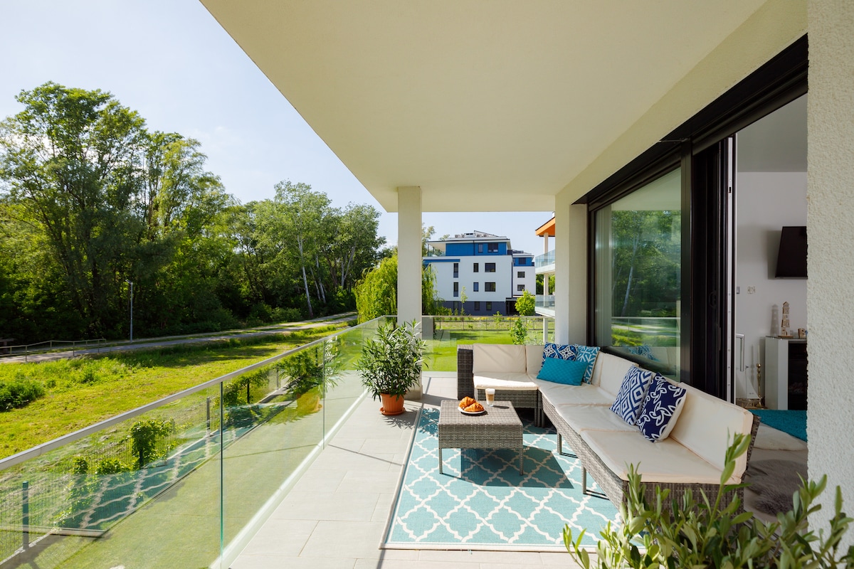 A spacious balcony features a comfortable seating area with patterned cushions and a table. Lush greenery is visible beyond the glass railing, and a colorful rug adds a touch of warmth. A serene atmosphere is created by the surrounding landscape.