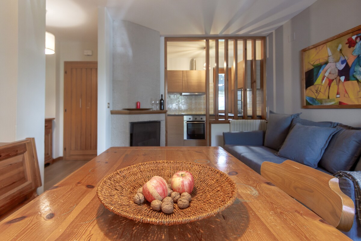 A dining area features a wooden table with a round woven bowl containing apples and walnuts. In the background, a cozy living space is visible, highlighted by a large painting and a modern kitchen area with integrated appliances. Natural light filters in through the windows.