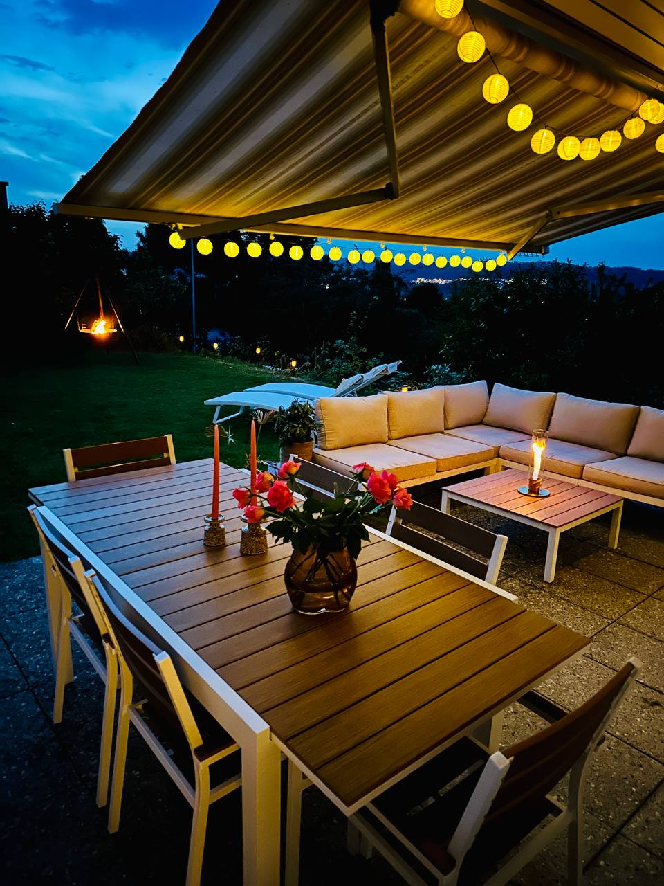 A covered outdoor seating area features a wooden table surrounded by white chairs, enhanced by a bouquet of flowers. A cozy sectional sofa and coffee tables provide additional seating. Soft string lights above create a warm ambiance, while a lantern adds a touch of elegance.