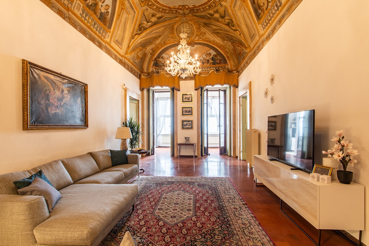 A spacious living area showcases a decorative ceiling adorned with historical frescoes. An elegant chandelier hangs overhead, illuminating the room. A large sectional sofa invites relaxation, while a flat-screen TV and decorative accents complement the stylish design. Windows provide ample natural light.