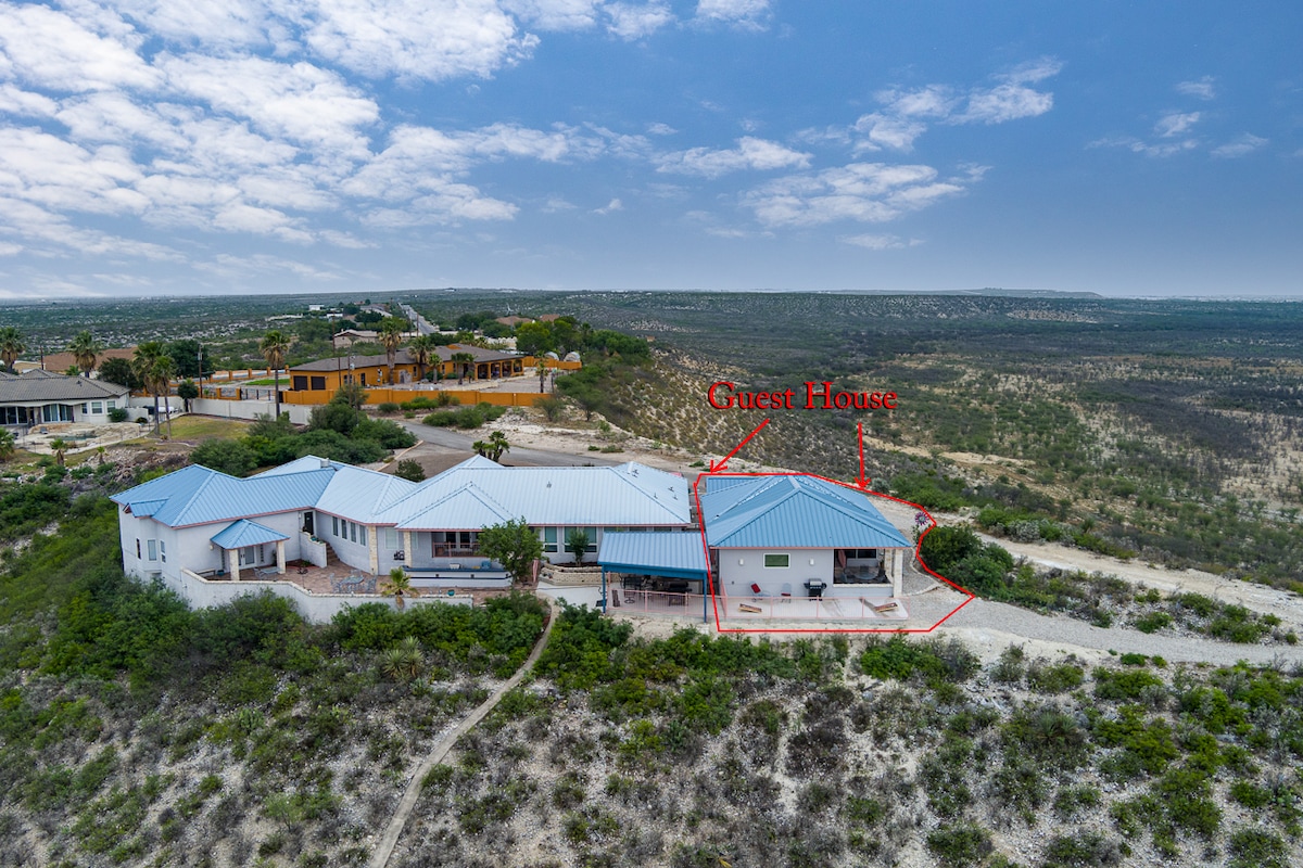 An aerial view displays the guest house amidst the natural landscape, featuring a distinctive blue roof. Surrounding greenery and unspoiled terrain highlight the home's location, while the structure's layout reveals multiple connected sections and a covered outdoor area.