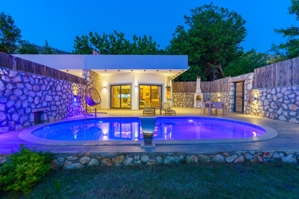 A tranquil outdoor area is highlighted by a swimming pool, which is illuminated in soft blue and surrounded by natural stone walls. Seats and a dining area are positioned near the pool, with a cozy hanging chair nearby, offering a relaxing spot to enjoy the surroundings.