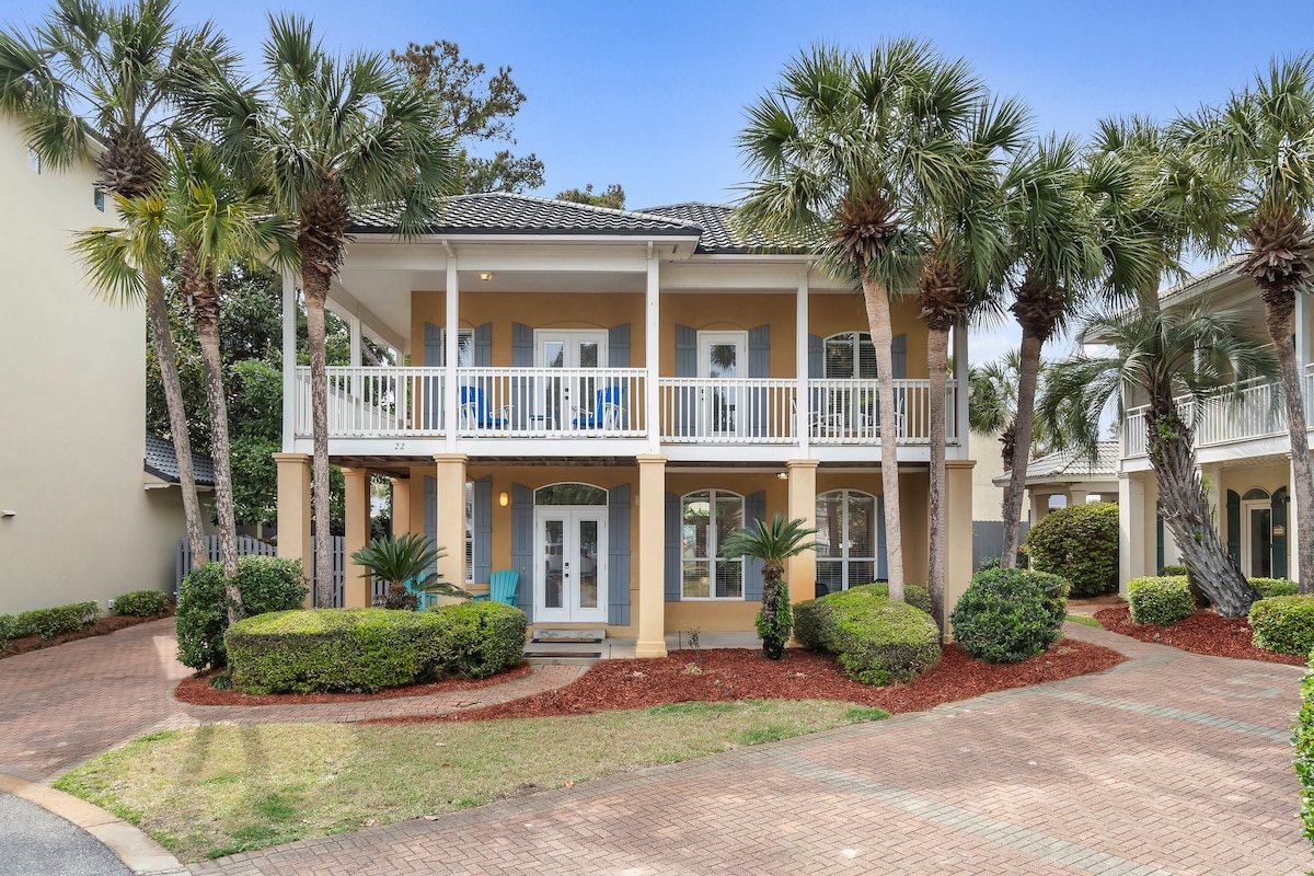 A luxury beach house is showcased with a welcoming façade, featuring a covered upper porch with blue chairs and palm trees lining the entrance. A well-maintained lawn surrounds the home, and a cobblestone driveway leads to the structure, framed by neatly trimmed shrubs.
