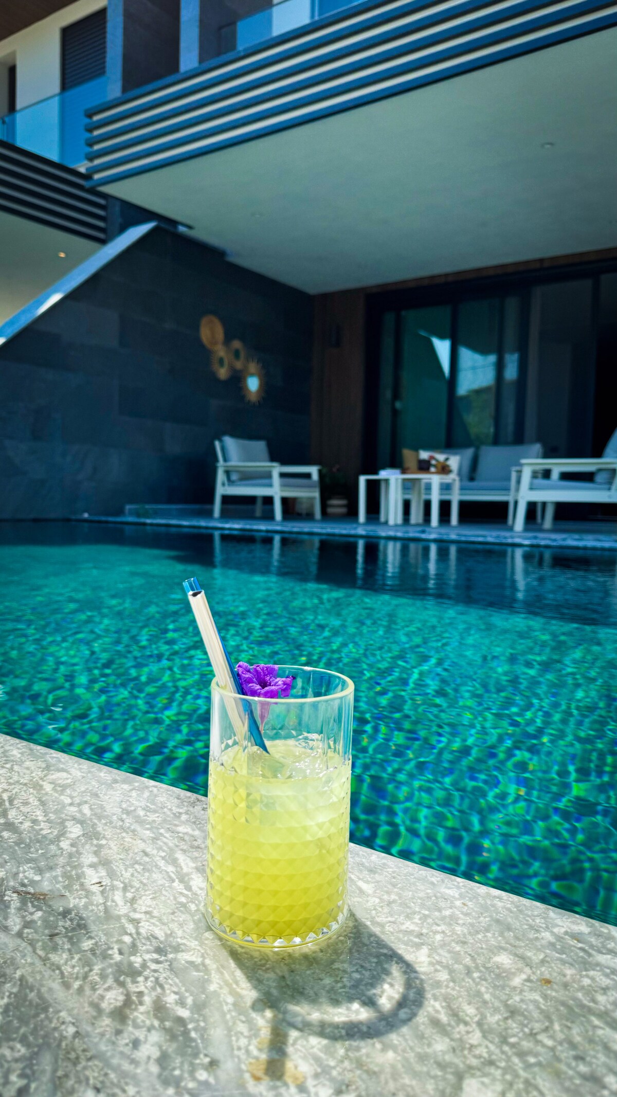 A glass of a vibrant yellow drink topped with a decorative flower is placed on a stone surface. In the background, an inviting swimming pool with clear, shimmering water reflects the modern villa featuring comfortable seating areas.