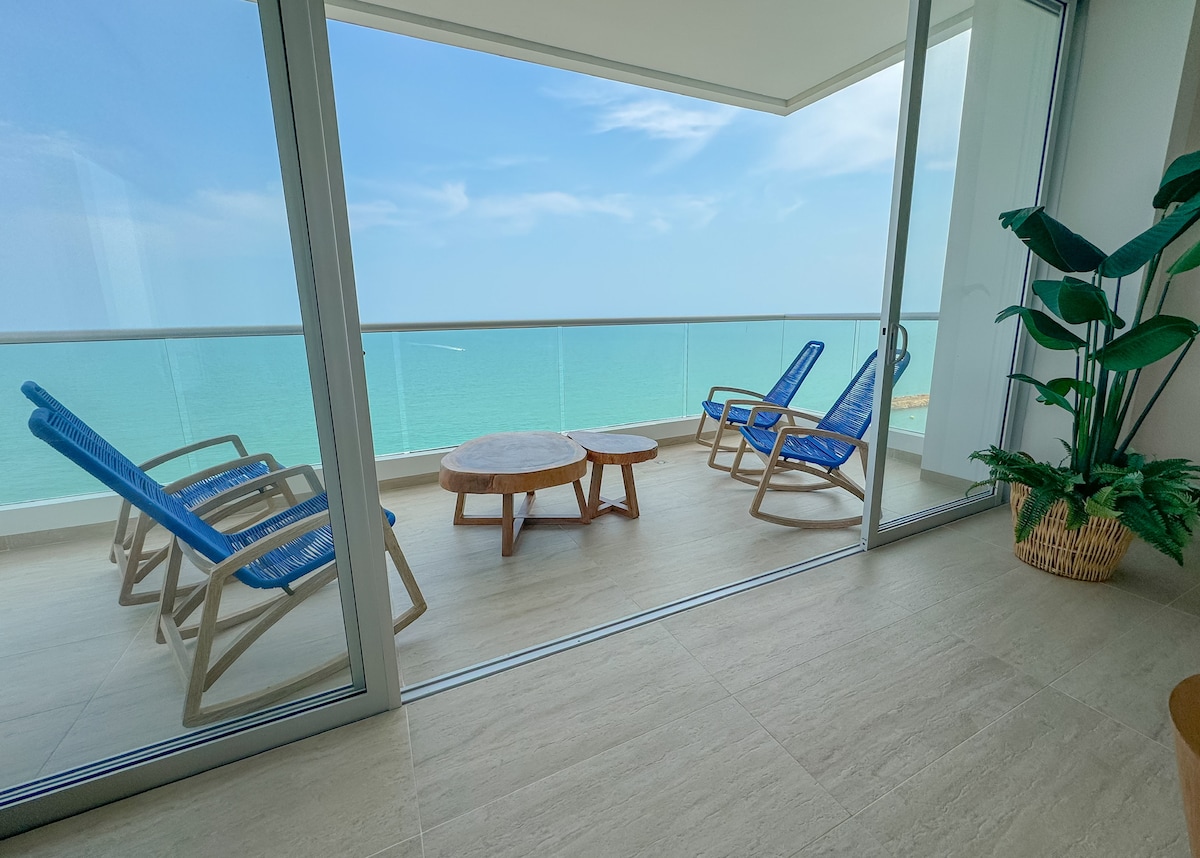 A balcony with panoramic sea views features two blue reclining chairs alongside a round wooden table. Natural light floods the area, and green foliage is visible in a nearby planter, creating a relaxing environment.