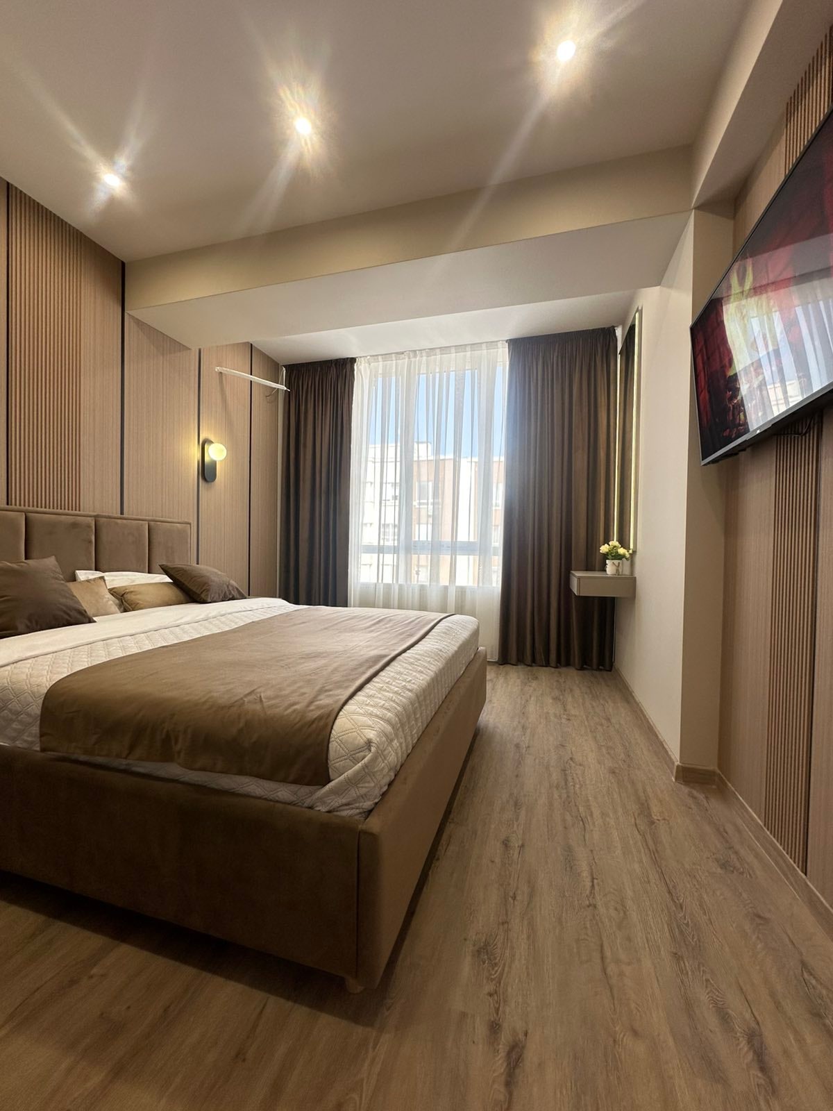 A stylish bedroom features warm, neutral tones with wooden flooring. A spacious bed is dressed in soft linens, complemented by decorative pillows. Natural light streams through large windows, adorned with dark curtains, and a flat-screen television is mounted on the wall opposite the bed.