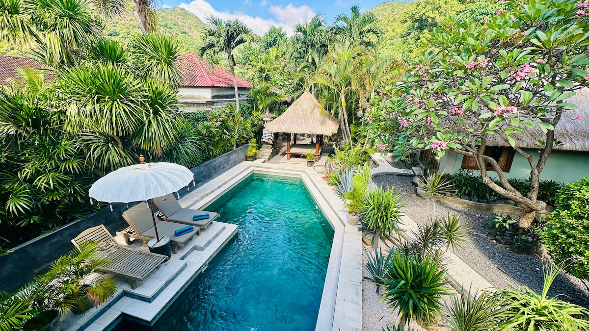 A crystal-clear pool is surrounded by lush tropical plants and trees, offering a serene atmosphere. A shaded wooden gazebo provides comfortable seating, while lounge chairs with an umbrella are positioned beside the water, inviting relaxation in the sun-drenched garden.