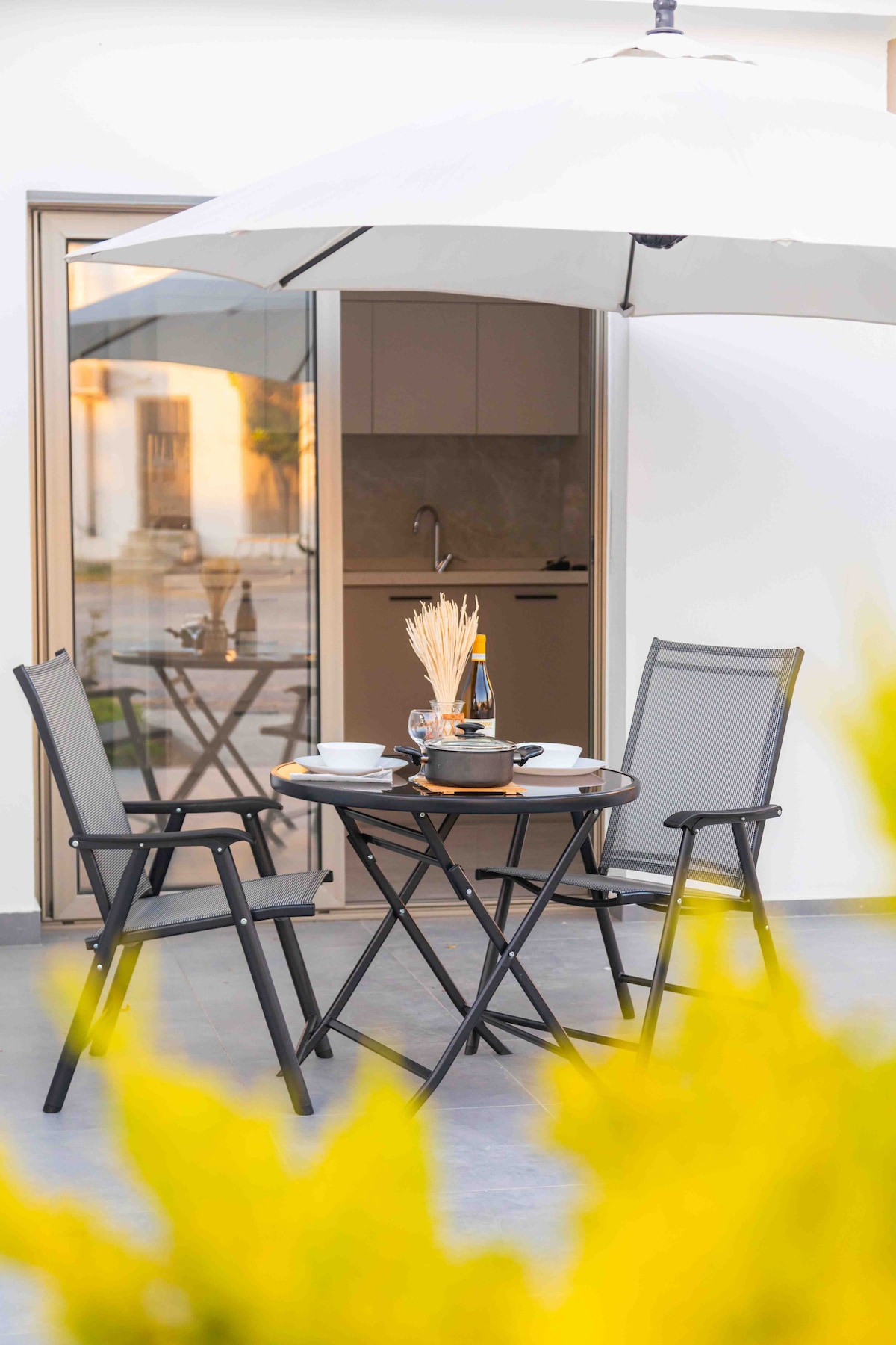 A private patio space is featured, furnished with a round table and two chairs under a large umbrella. The scene includes a selection of tableware arranged on the table. In the background, a kitchen area is visible through glass doors, merging indoor and outdoor living.