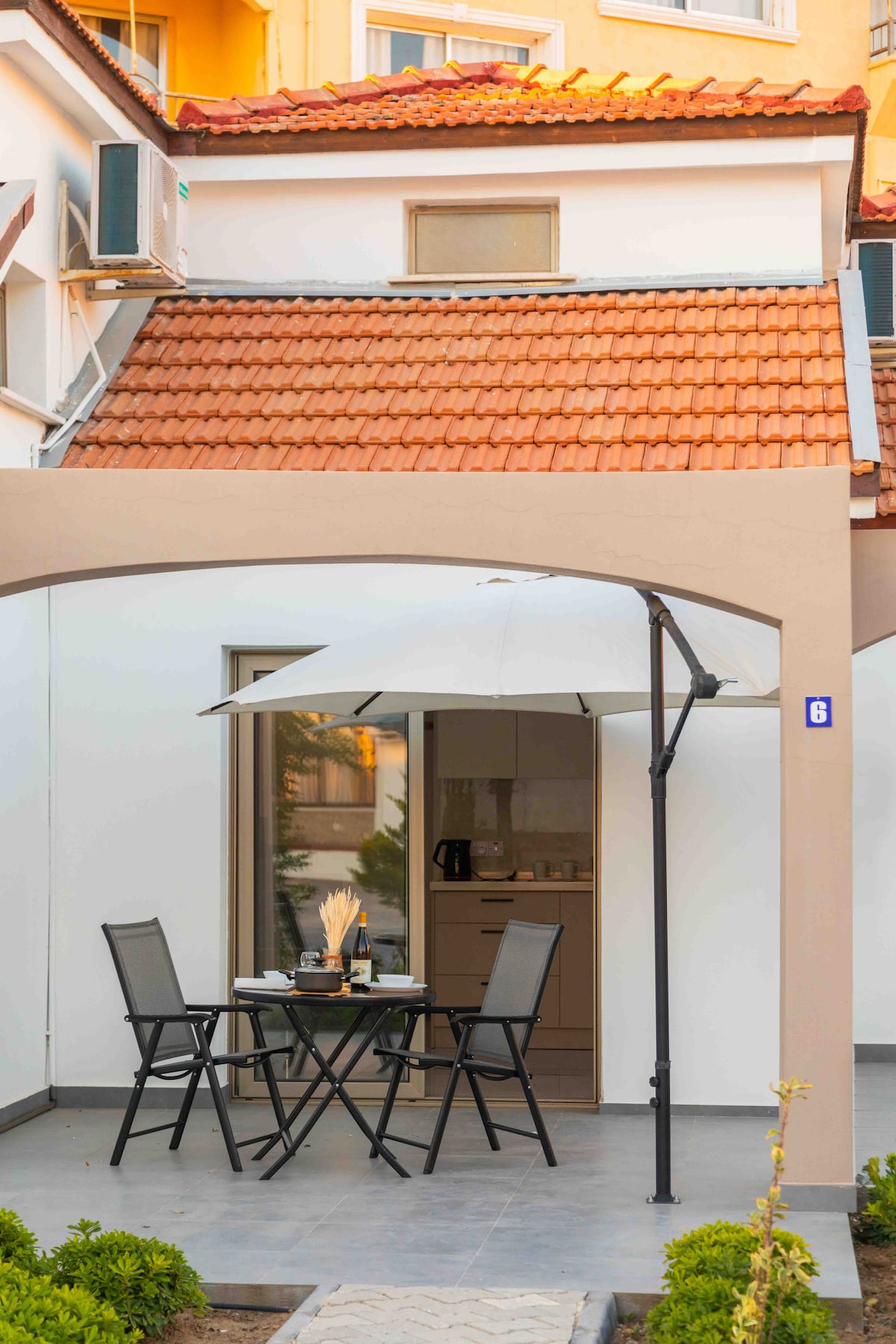 A private patio is featured, furnished with a small round table and four black folding chairs. A white umbrella provides shade, creating a comfortable outdoor space. The entrance to the bungalow is visible through a glass door, leading to the interior.