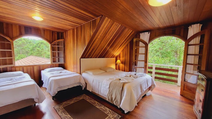 A cozy guest room features three beds, including a spacious bed and two smaller beds. Natural wood paneling creates a warm atmosphere. Large arched windows offer views of the surrounding greenery, while soft lighting enhances the inviting environment.