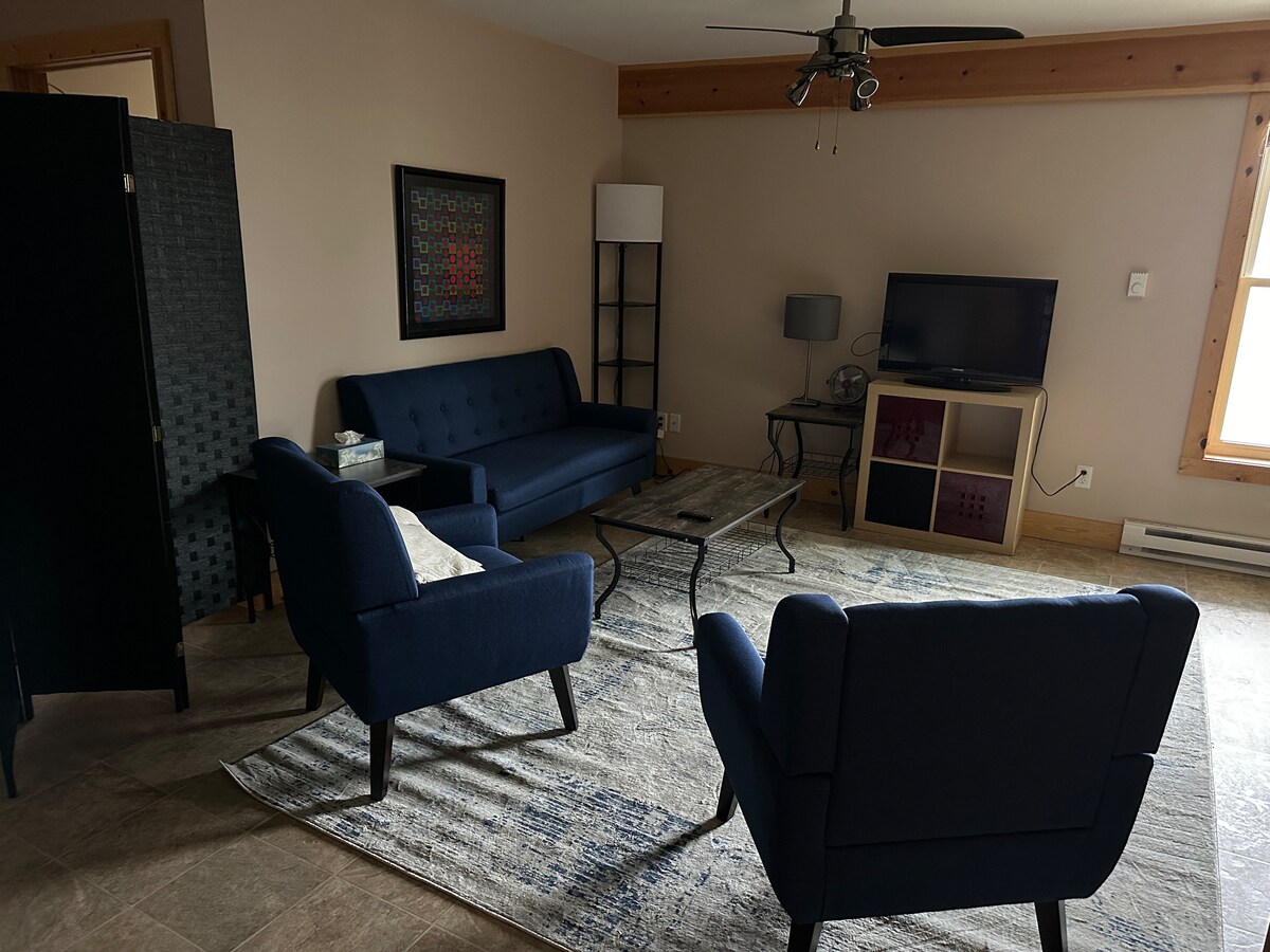 A living area features a navy blue sofa alongside two matching armchairs, set on a textured light-colored rug. A glass coffee table sits in the center, while a TV stand with a television and a lamp is positioned nearby. Natural light enters through a window.