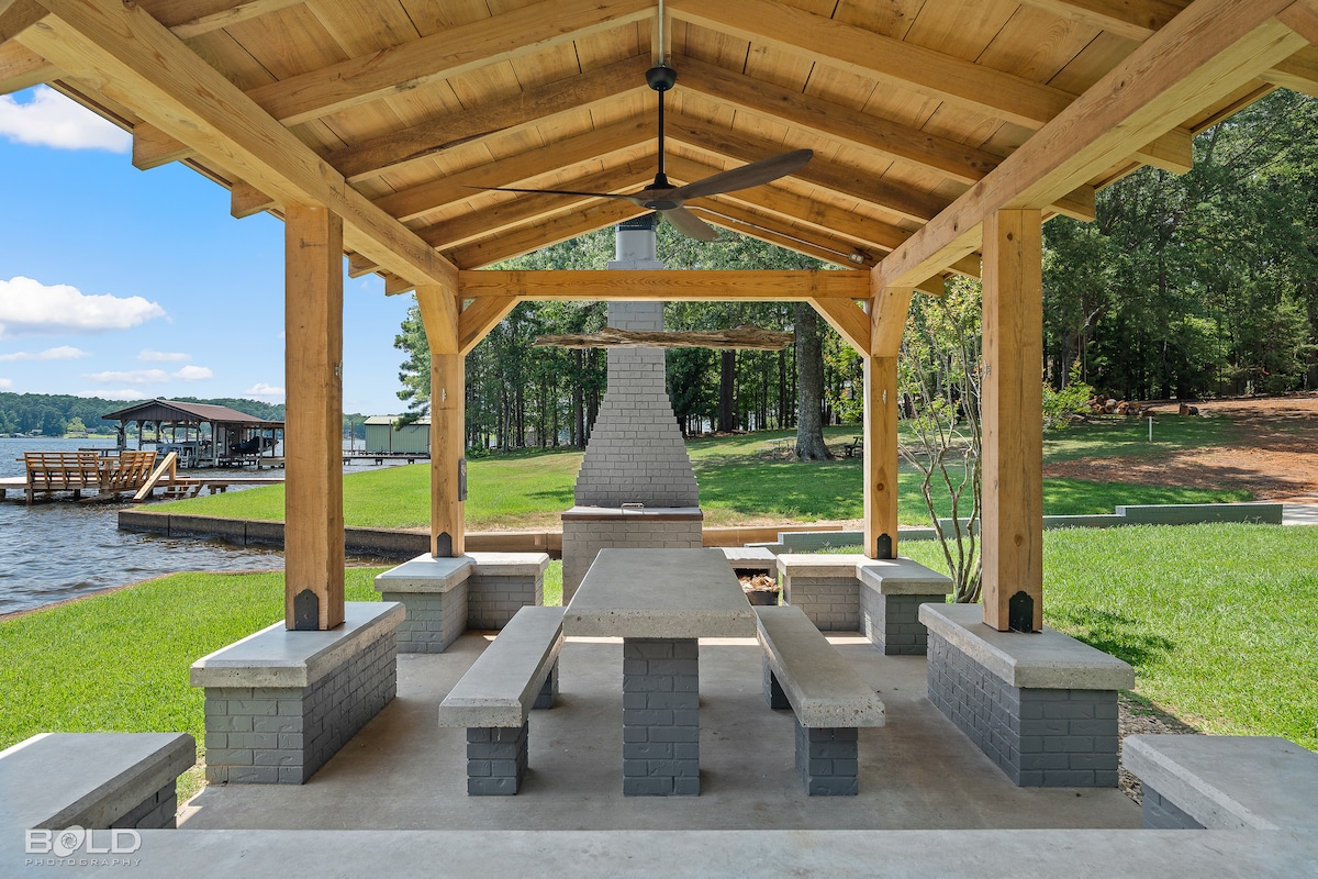 A spacious picnic pavilion is shown, featuring a large concrete table surrounded by stone benches. The structure is covered by a wooden roof supported by sturdy beams. A stone fireplace is positioned at the back, with a view of the lake and green grass framing the scene.