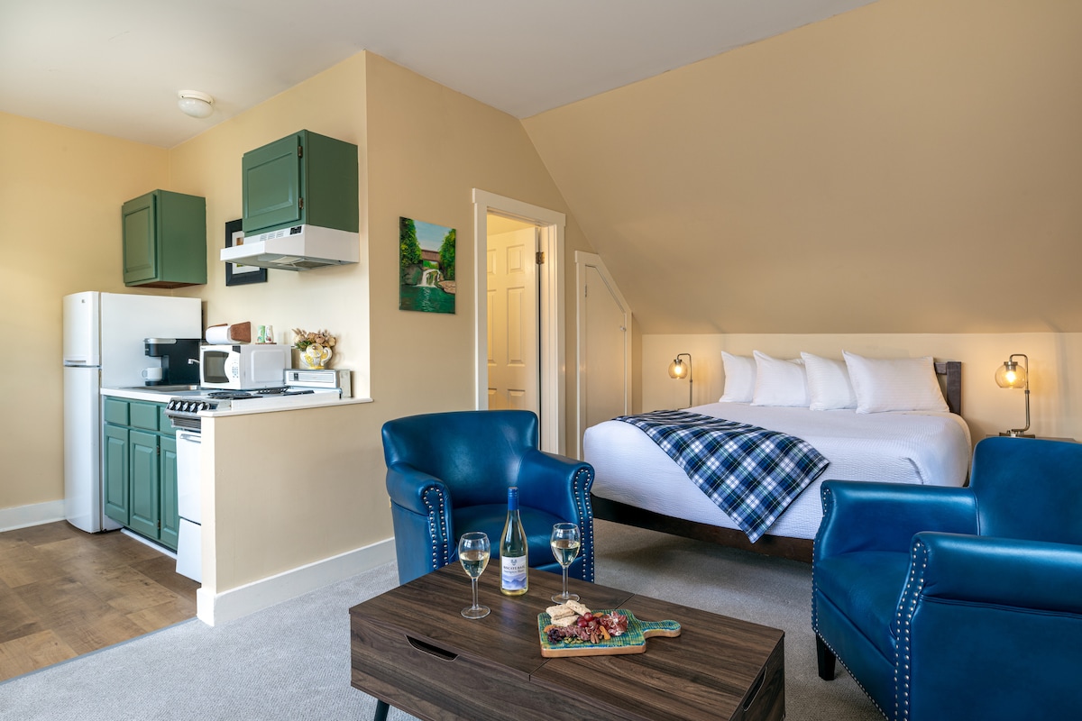 A bright studio suite features a comfortable sleeping area with a spacious bed dressed in soft linens. A kitchenette displays modern amenities, including a refrigerator and microwave. Two blue armchairs are positioned around a wooden coffee table, presenting a cozy space for dining or relaxation.