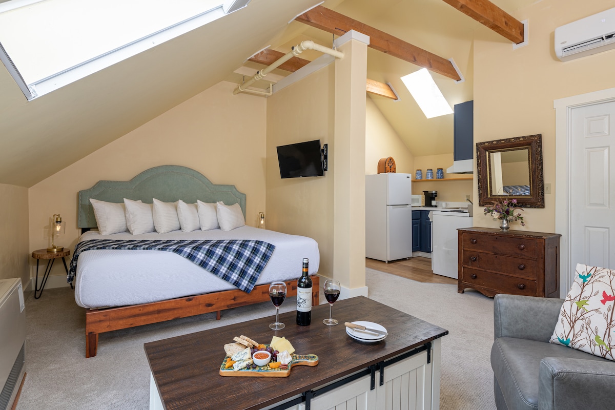 A spacious suite is featured, showcasing a king-size bed with multiple pillows arranged against a soft-colored headboard. Skylights enhance natural light, and a small kitchenette is visible in the background. A cozy seating area includes a gray chair, while a coffee table is set with wine and a snack.