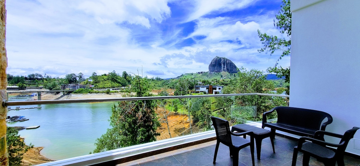 A spacious balcony features outdoor seating with two chairs and a small table. Scenic views of a shimmering lake and lush greenery are complemented by the striking silhouette of the Piedra del Peñol in the background, under a bright and expansive sky.