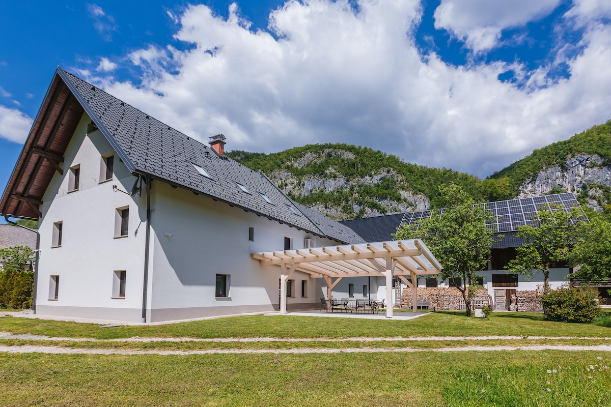 The exterior of a large, renovated farm building is showcased against a backdrop of mountains and blue sky. A covered terrace with outdoor seating is visible in the foreground, surrounded by well-maintained grass and greenery.