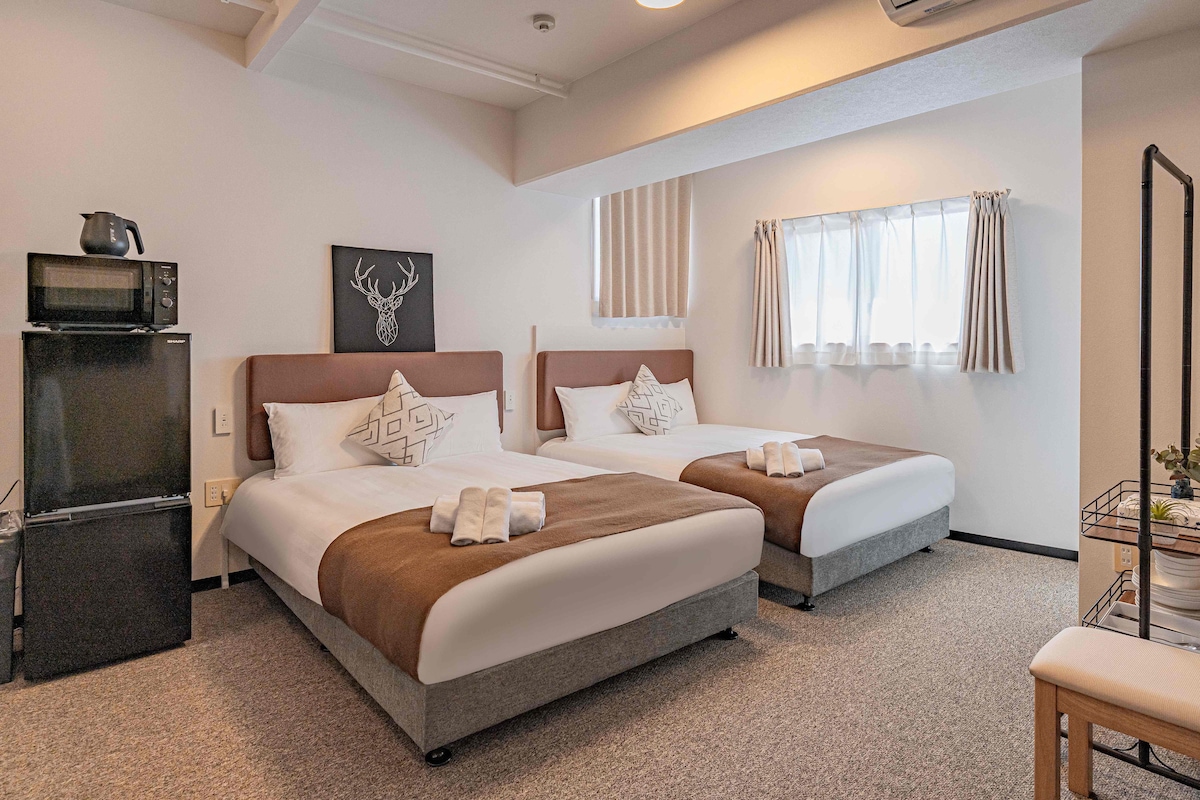 A spacious room is furnished with two beds, each topped with a decorative pillow and a set of fresh towels. A small refrigerator is positioned in the corner next to a kettle. Natural light enters through a window dressed with light curtains.