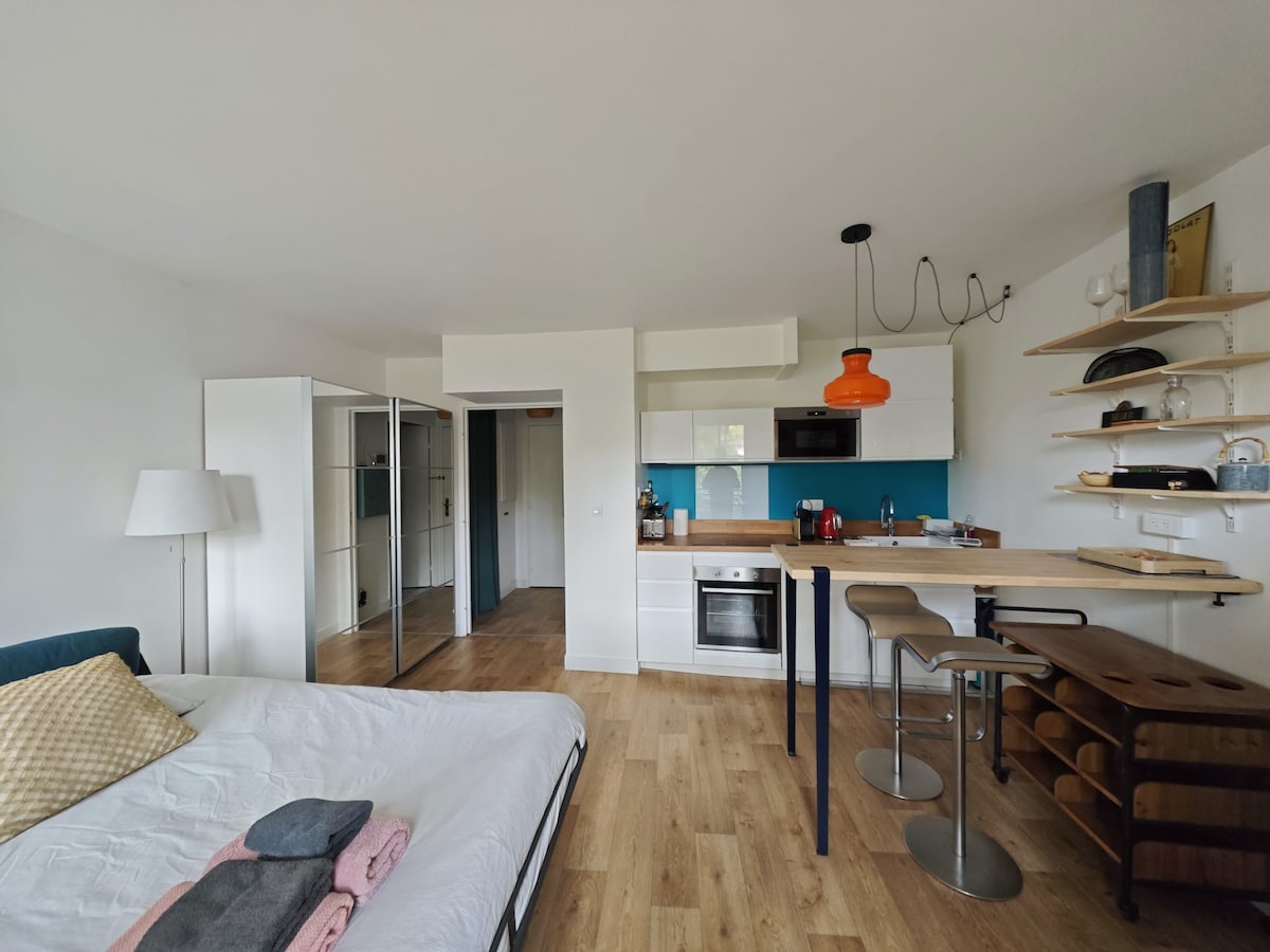 A bright studio space features a spacious bed with neatly arranged blankets, a modern kitchenette equipped with essential appliances, and a dining area with two stools. Natural light flows through large windows, and stylish shelving displays various items, emphasizing a contemporary design.