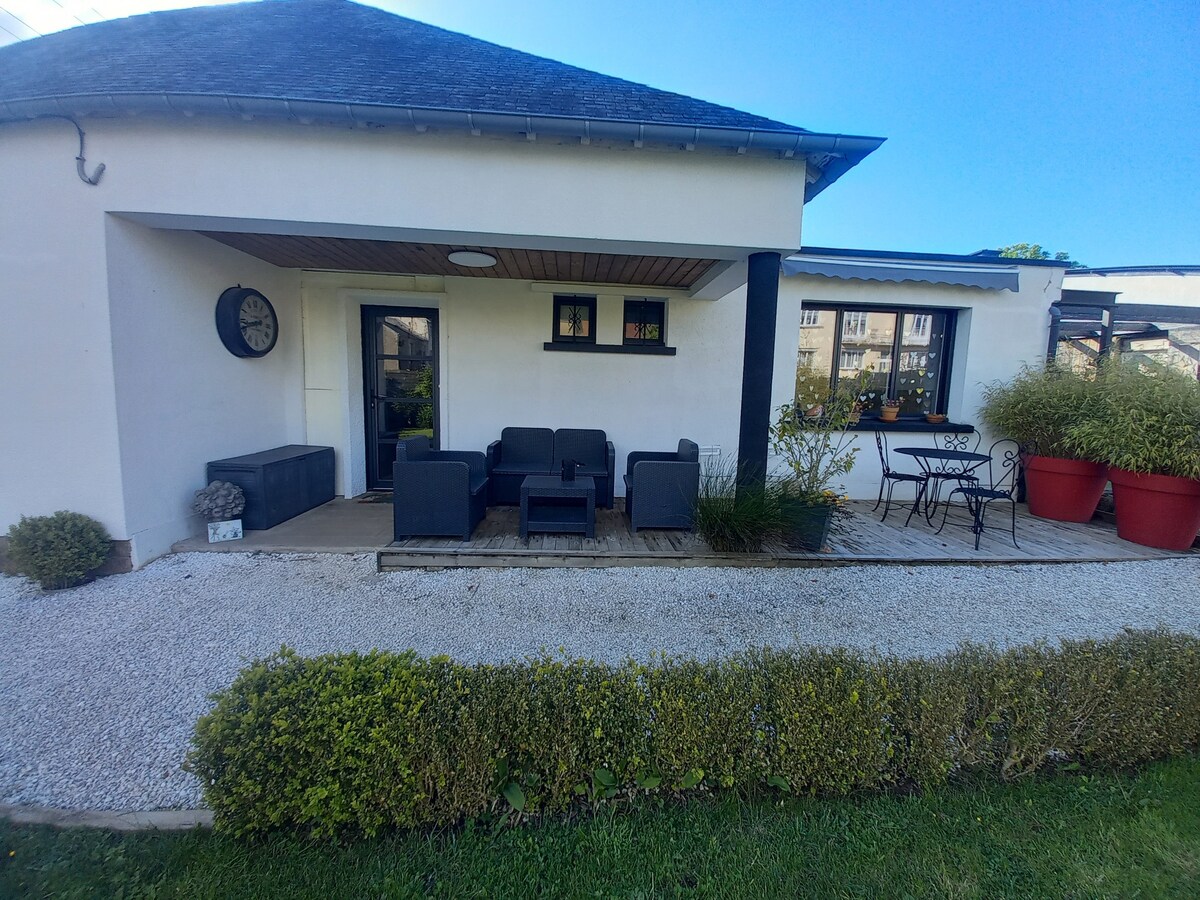 An outdoor seating area features a dark lounge set on a wooden terrace, framed by low greenery and flower pots. A small table with chairs is positioned beside the entrance, while a clock adorns the wall, adding functionality to the serene space.