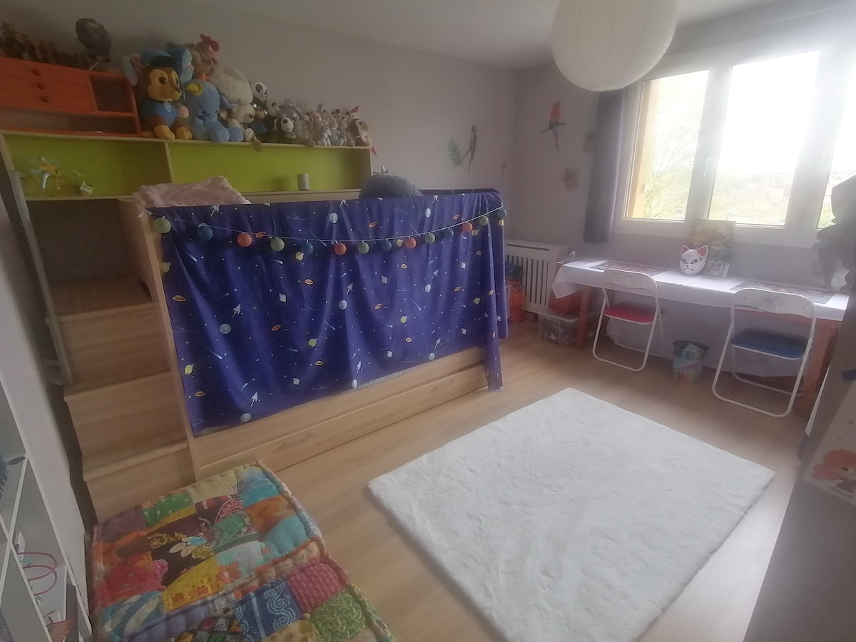 A children's bedroom features a soft, light-colored rug on wooden flooring. A playful bed area is enclosed with a colorful curtain, while a small display of stuffed animals is arranged above. Two desks with chairs are positioned by the window, providing a well-lit space for activities.