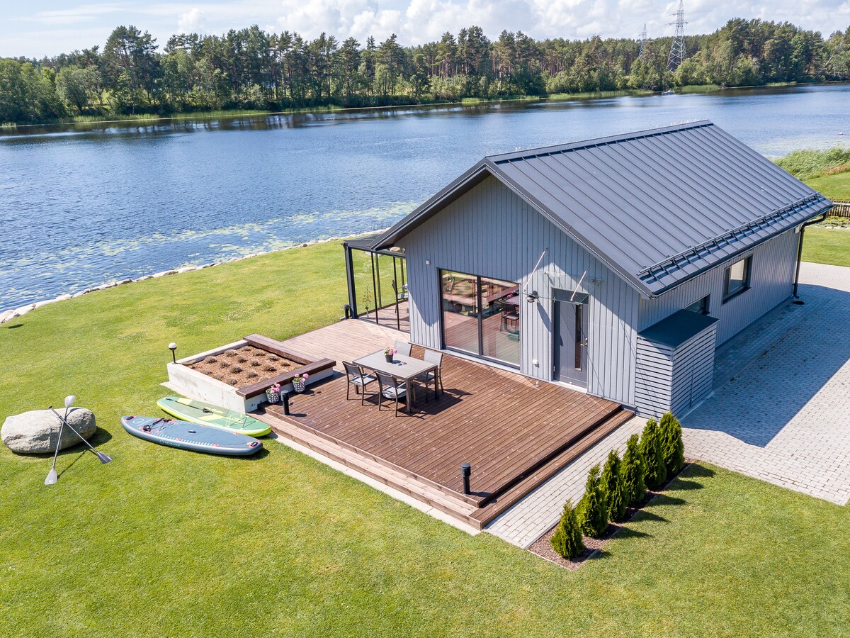 The modern villa is set beside a calm river, featuring a spacious wooden deck with outdoor dining furniture. Two paddleboards and a kayak rest nearby. The surrounding lush green lawn and trees provide a serene backdrop, complemented by large glass doors inviting natural light into the interior.