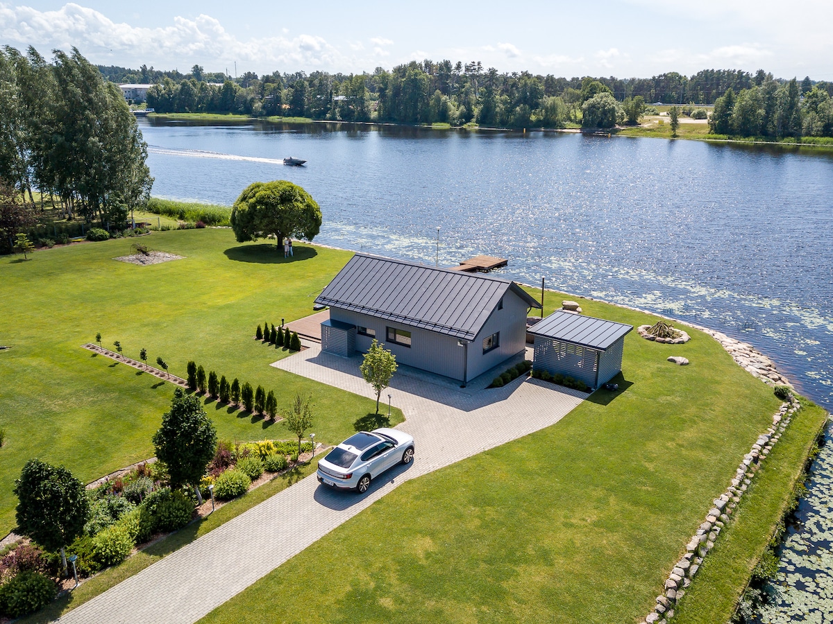 The villa is situated beside the tranquil waters of the Pärnu River, surrounded by a grassy area and landscaped gardens. A paved driveway leads to the entrance, with a car parked outside. Lush greenery adds to the serene setting, enhancing guest relaxation.
