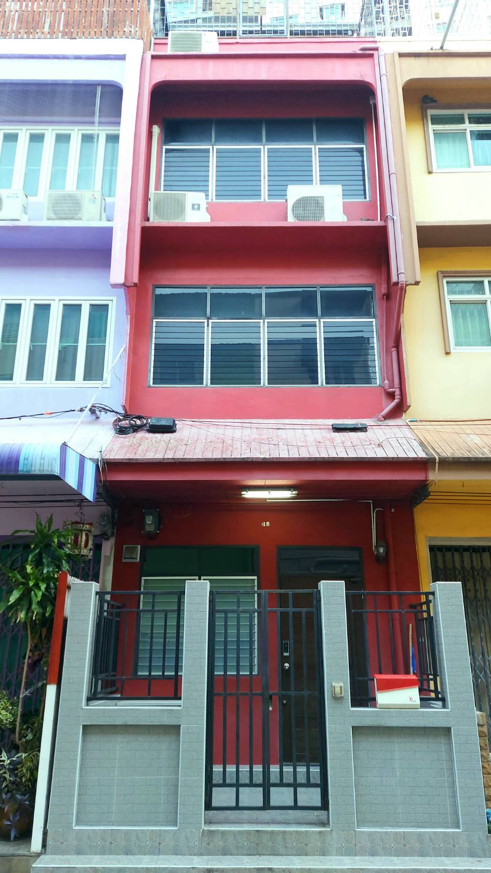 A three-story house with a vibrant red facade is presented. The front entrance is secured with a black gate, and the ground level features a neutral-toned wall. Air conditioning units are positioned on the upper floors, while colorful neighboring buildings enhance the inviting street view.