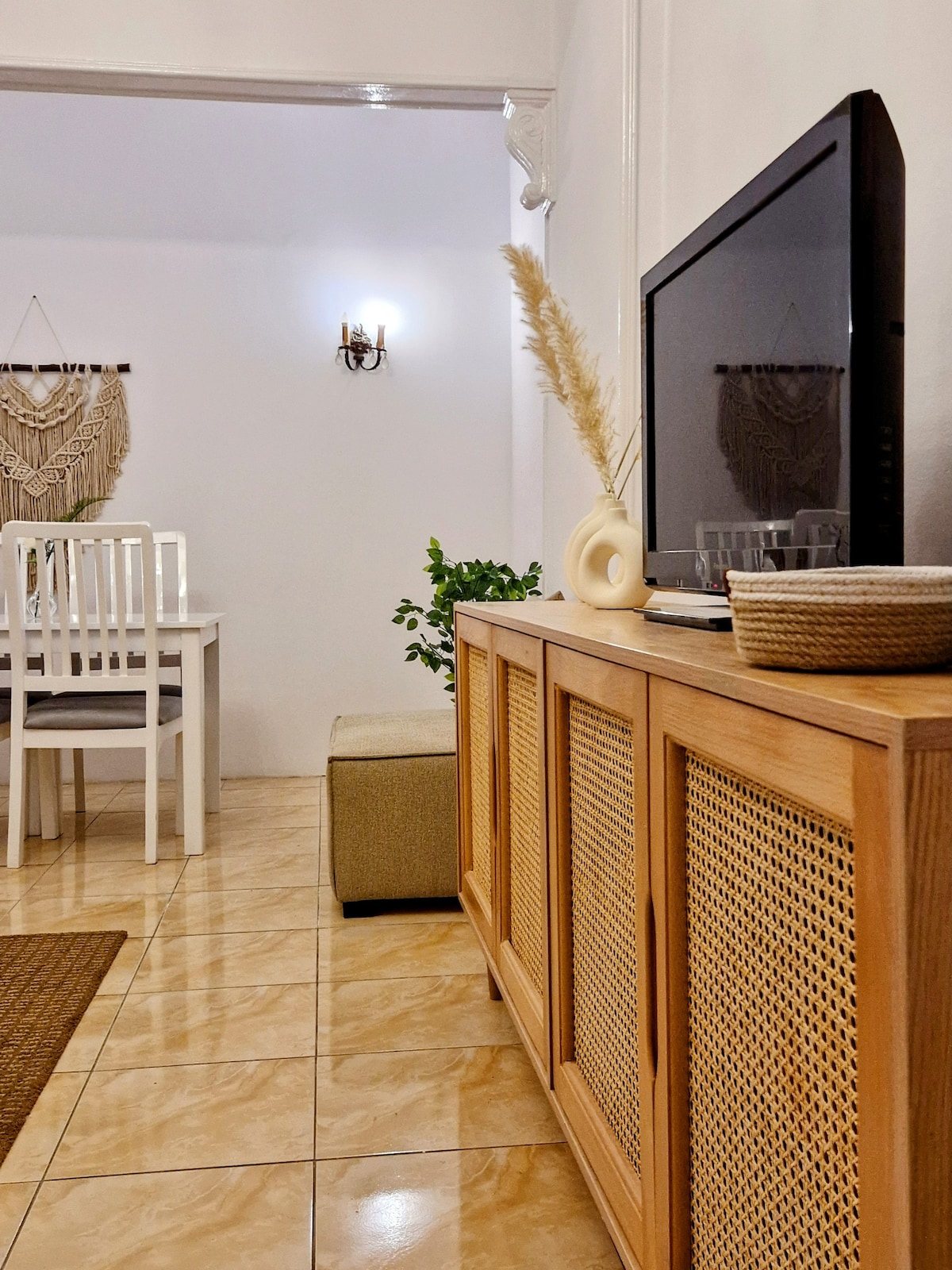 A clean and modern living area is shown, featuring a wooden media console with decorative accents and a television. A dining table with white chairs is visible in the background, along with a cozy seating area. Soft lighting adds to the space's welcoming feel.