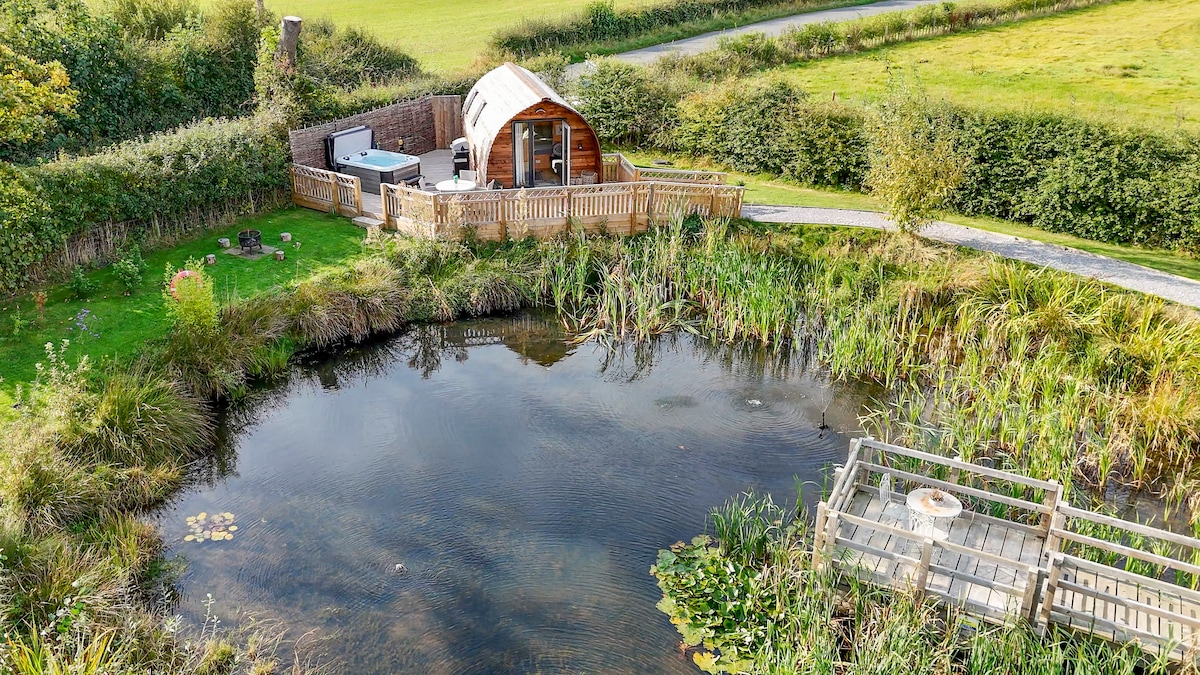 A scenic view of a glamping pod situated beside a tranquil pond, surrounded by lush greenery. The pod features a spacious deck with a hot tub and seating area. A wooden path leads toward the pond, enhancing the serene countryside atmosphere.