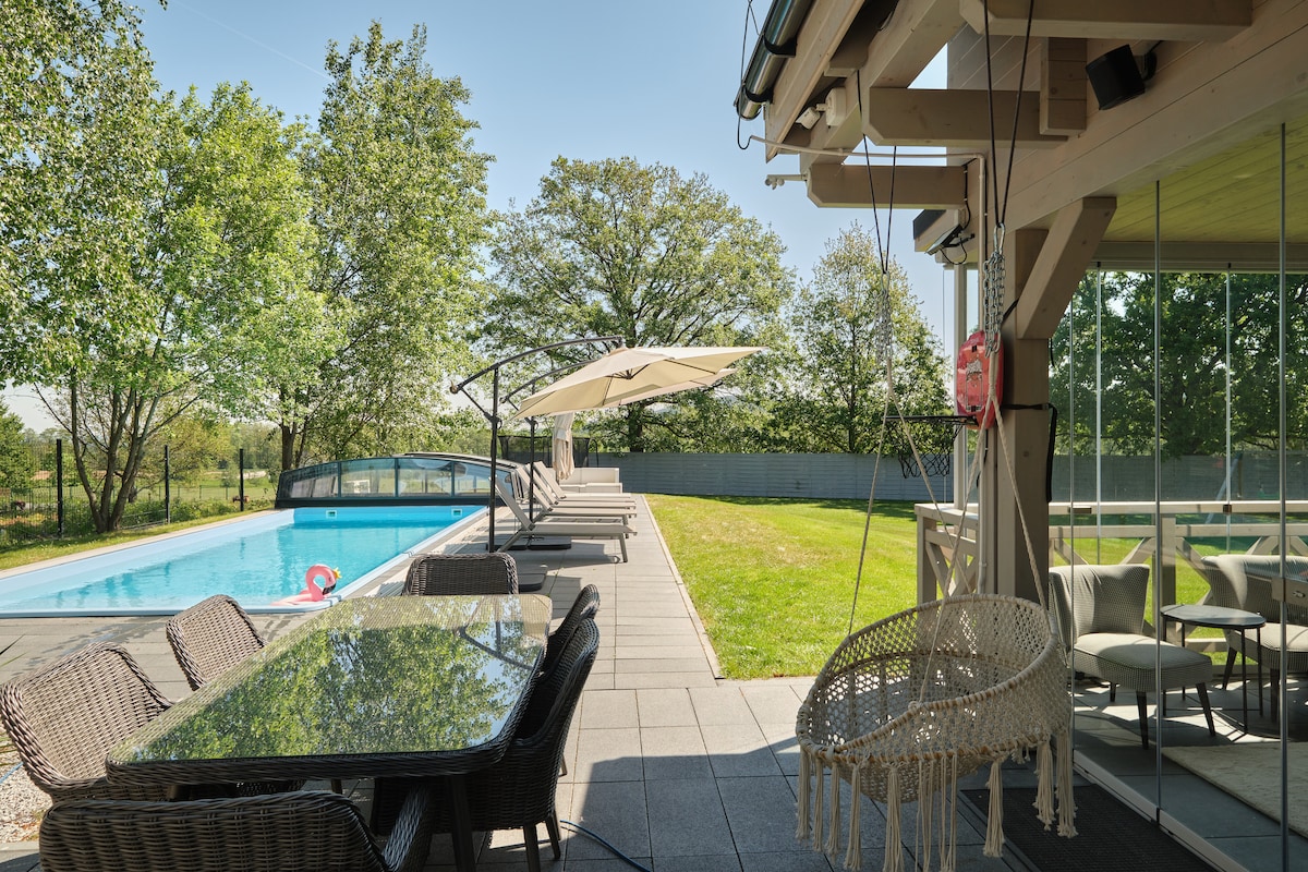 A spacious outdoor area features a well-maintained pool surrounded by sun loungers and umbrellas. A large dining table is positioned near a glass-walled terrace, providing a view of the manicured lawn and trees offering shade in the background.