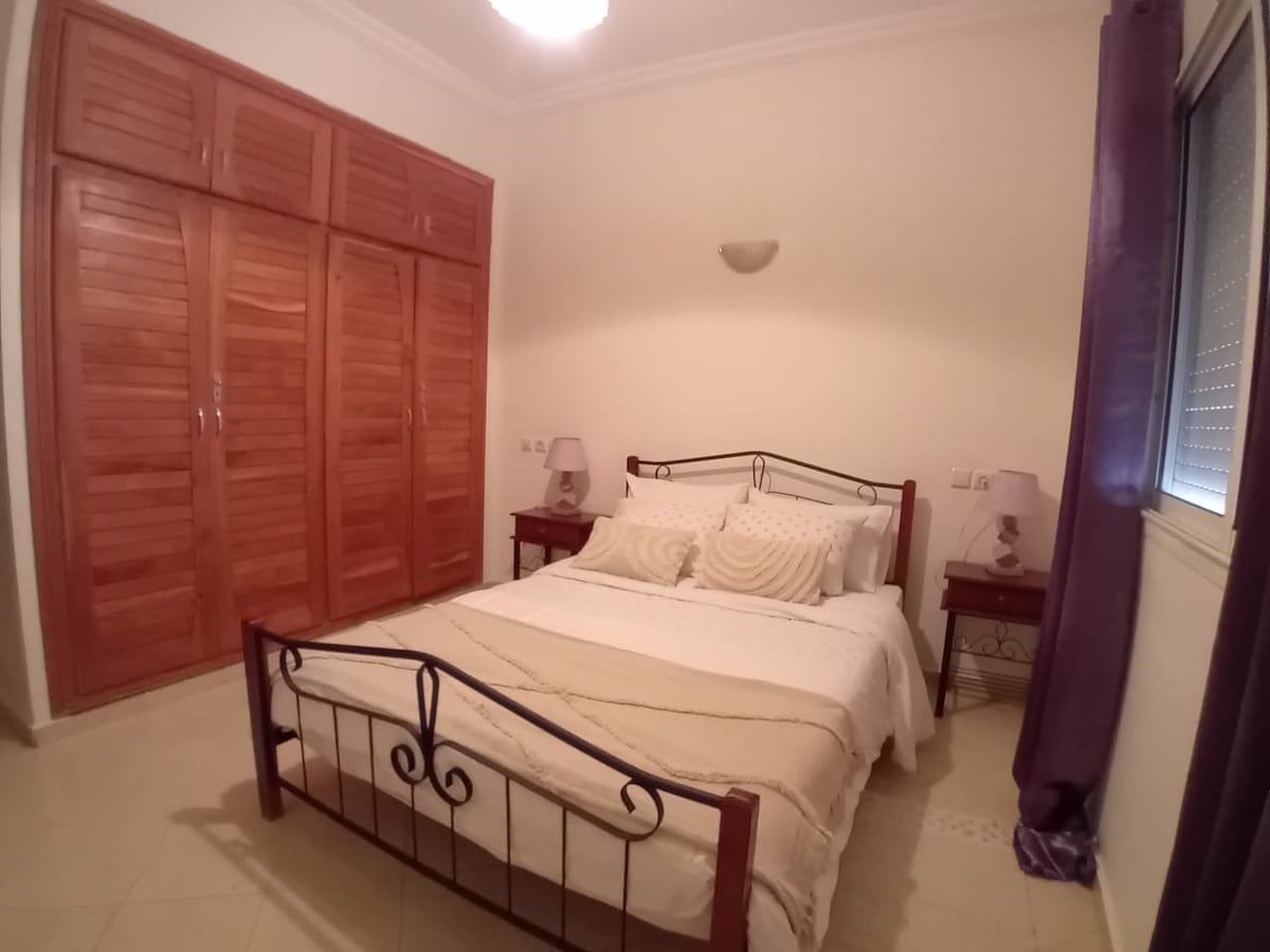 The bedroom features a spacious bed adorned with neutral bedding, complemented by two bedside tables each with lamps. A large wooden wardrobe spans one wall, while light walls enhance the room's brightness. Natural light filters through the window, which is covered by simple blinds.