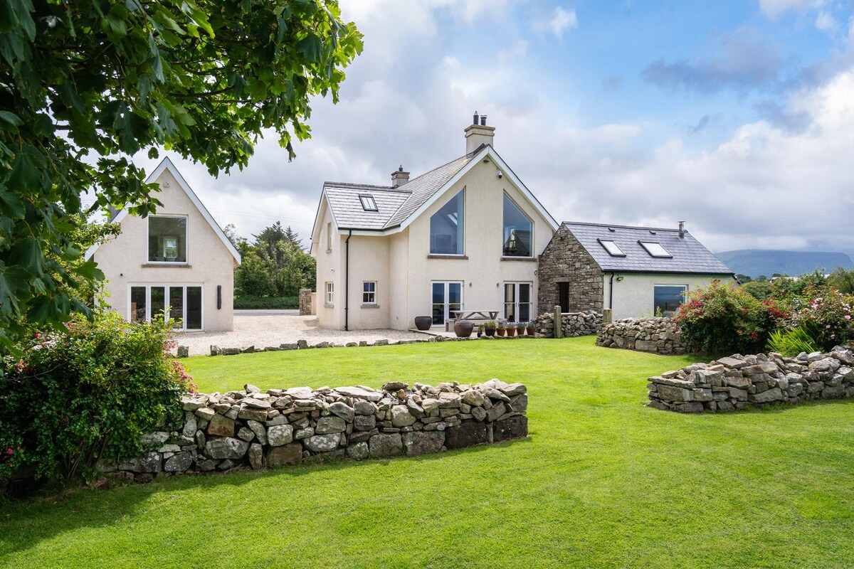 A serene exterior view of a multi-level house surrounded by lush greenery. Stone walls and landscaped gardens add character, while large windows reflect natural light. Two distinct architectural styles are visible, with a blend of traditional stone and modern design elements.