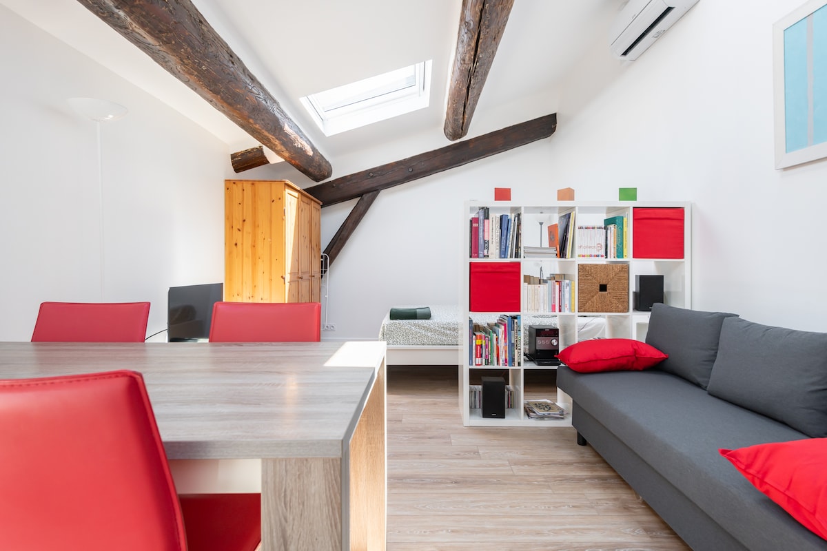 The living space features exposed wooden beams and a skylight, providing ample natural light. A gray sofa with red cushions is positioned near a bookshelf filled with books. A dining table with red chairs is visible, along with a neatly made bed in the corner.