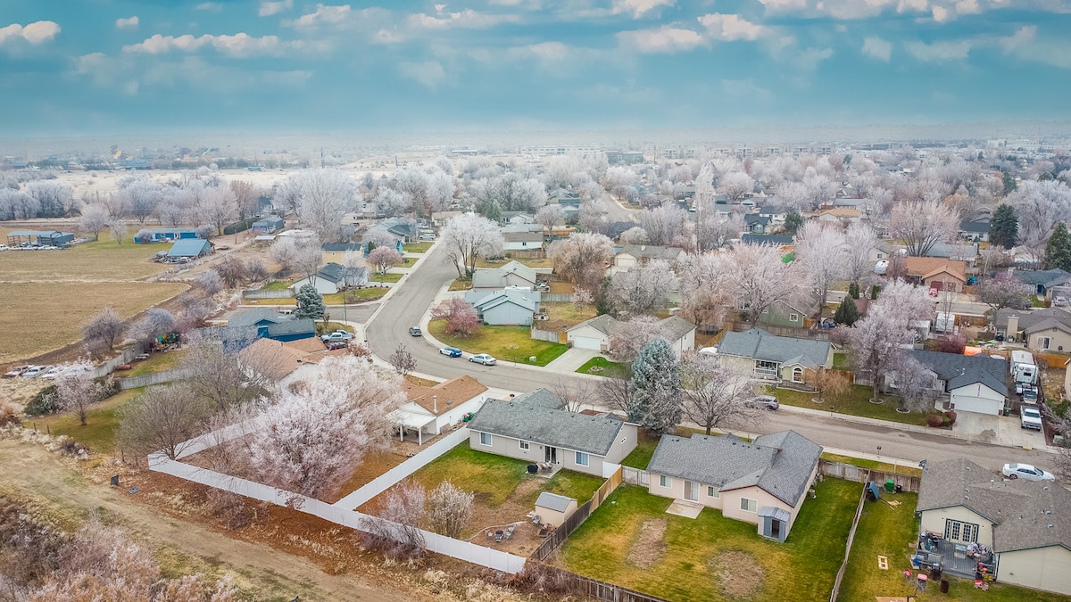 An aerial view captures a quiet residential neighborhood with trees adorned in frost. Well-kept homes are lined along the streets, with a large backyard visible in the foreground, providing a sense of spaciousness. Open fields extend beyond the edges of the community.