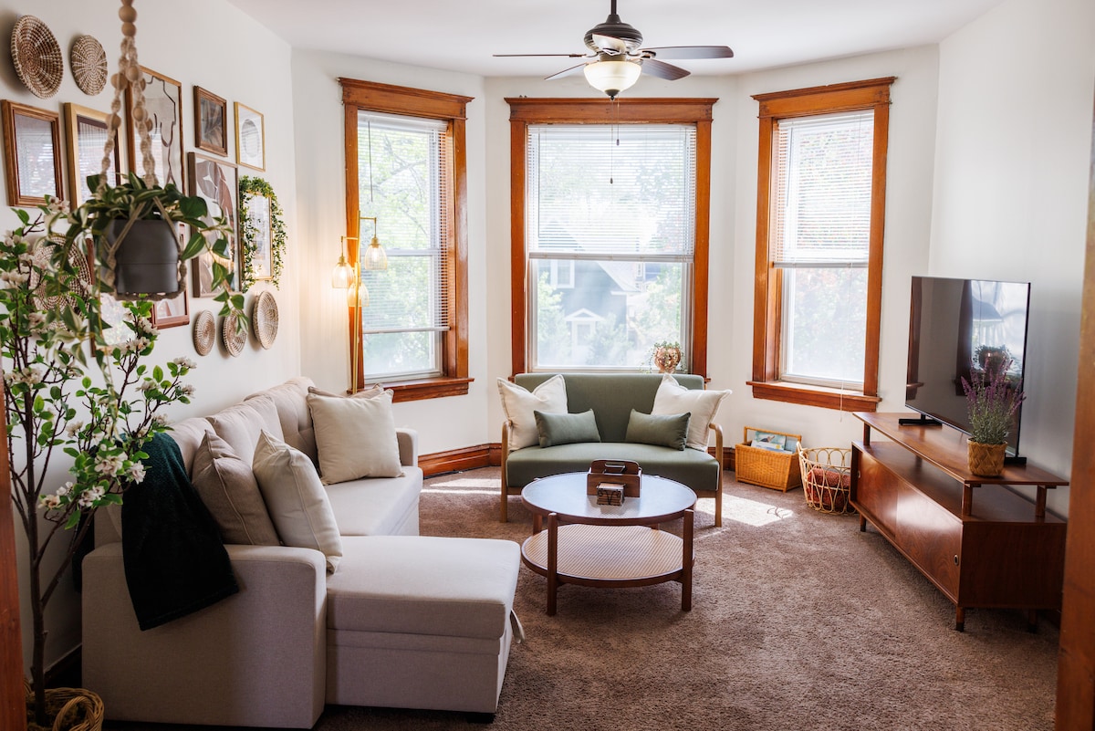 The living room features large windows allowing natural light to fill the space. A comfortable L-shaped sofa and a green accent sofa are arranged around a wooden coffee table. Decorative wall art and plants enhance the inviting ambiance, along with a TV and wooden media console.