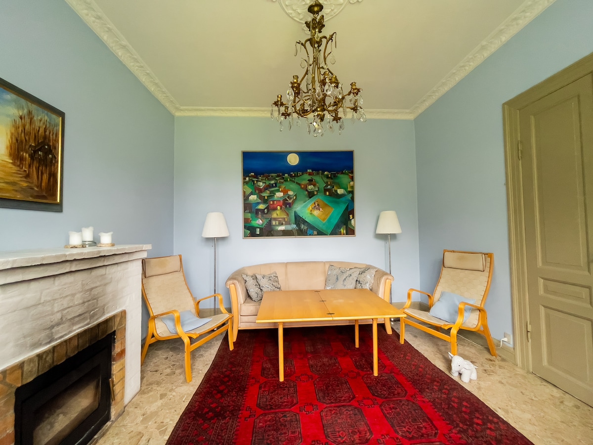 A living room is presented with a light blue color scheme. A central coffee table is surrounded by two wooden chairs and a light sofa. A large painting depicting a village scene hangs above a stone fireplace, and a chandelier adds overhead illumination.